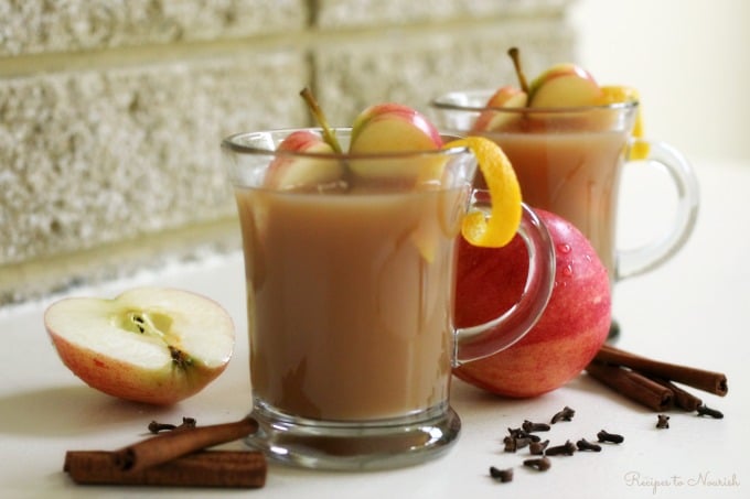 Glass mugs full of homemade apple cider with apple slices, orange zest, cinnamon sticks and cloves.