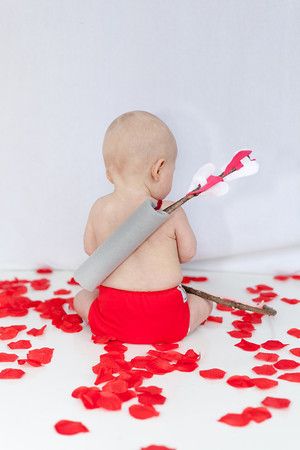 Baby sitting on floor surrounded by rose petals with fake quiver and arrows on their back