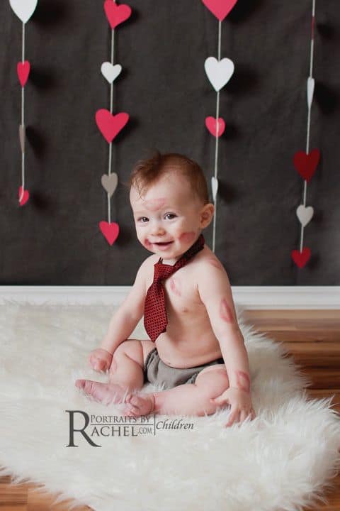 Baby sitting on floor with heart decor in the background and kiss marks on their face and arms 