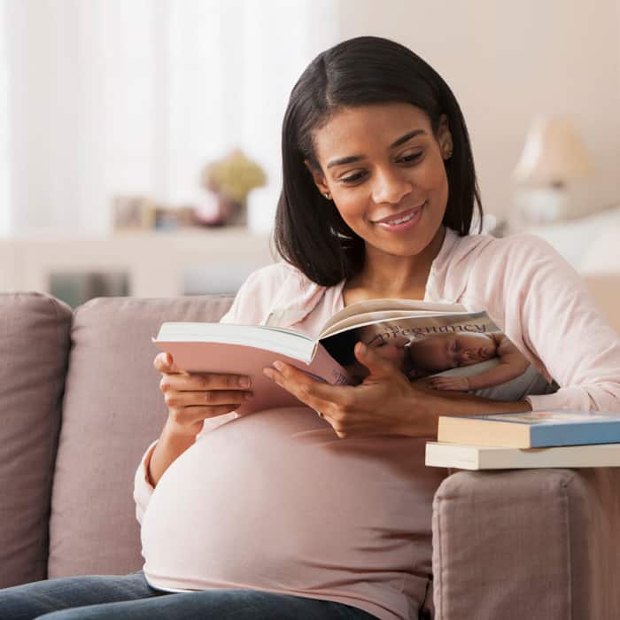 Pregnant woman reading stack of pregnancy books on her couch