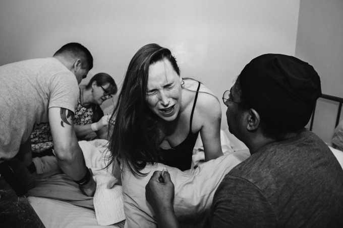 A mother in pain in active labor on her bed during a home birth surrounded by her husband and birth team.