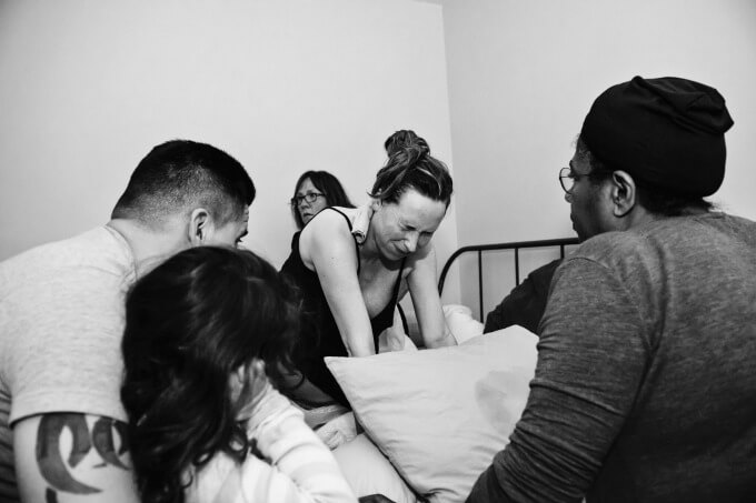 A mother in pain in active labor on her bed during a home birth surrounded by her husband and birth team.