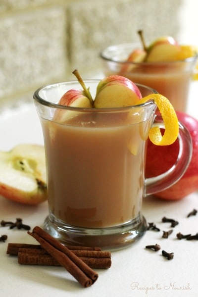 Glass mugs full of homemade apple cider, slices apples, orange zest, cinnamon sticks and cloves.