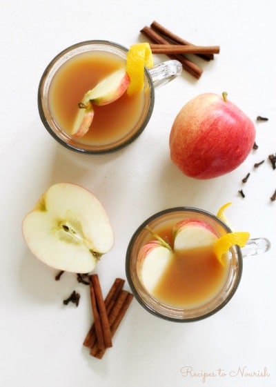 Glass mugs full of apple cider with apple slices, orange zest, cinnamon sticks and cloves.